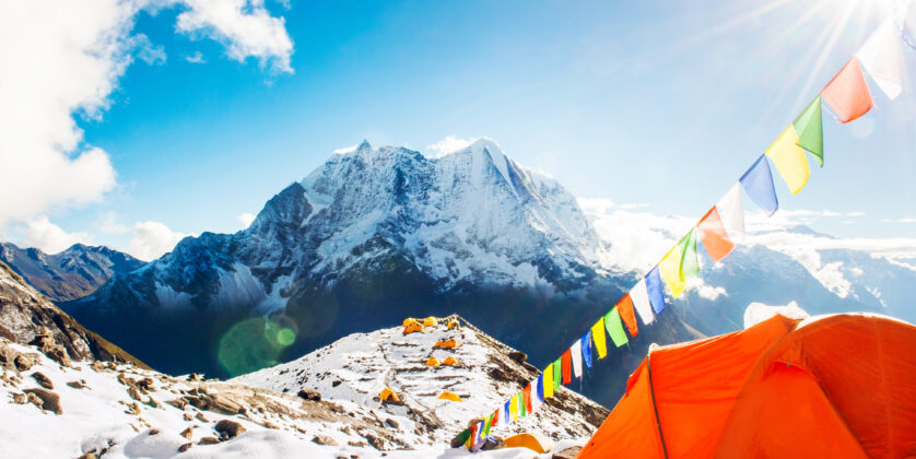 "A panoramic view of Everest Base Camp with snow-covered peaks, colorful prayer flags fluttering in the wind, and trekkers walking along the rocky terrain. In the background, Mount Everest towers majestically above the surrounding mountains under a clear blue sky."