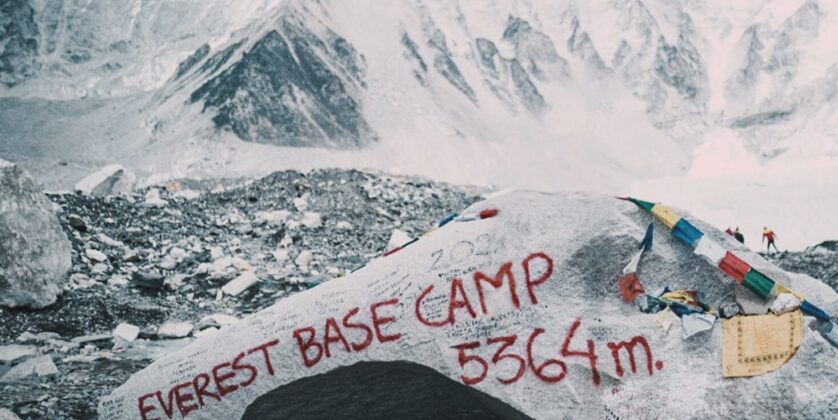 A panoramic view of Everest Base Camp at 5364 meters, showing clusters of colorful tents on a rocky, glacial morraine with majestic snow-capped Himalayan mountains in the background under a clear sky.