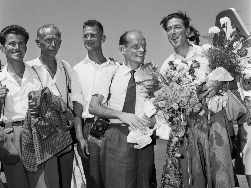 Group of male mountaineers standing on an airfield in 1956, smiling and holding large bouquets of flowers, photographed beside an aircraft after a Himalayan expedition.