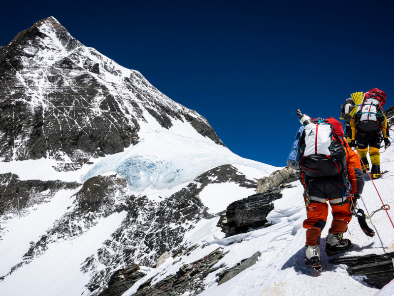 Climbers ascending the Geneva Spur buttress on Mount Everest, carrying expedition gear along a steep snow and rock ridge under a deep blue Himalayan sky.