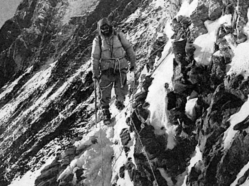 A climber from the 1956 Swiss Lhotse Expedition carefully traversing the steep Yellow Band at approximately 24,600 feet, using ropes and ice axes for support on the icy rock face.
