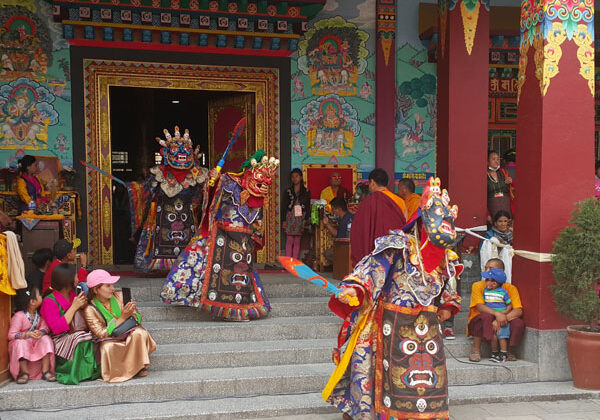 During the Mani Rimdu festival, dancers perform the traditional Chhingpa dance. The performers are dressed in elaborate, colorful costumes and intricate masks, representing various deities and mythical figures. The crowd watches from the steps and surrounding area outside a monastery as the dancers move energetically, invoking blessings and protection.