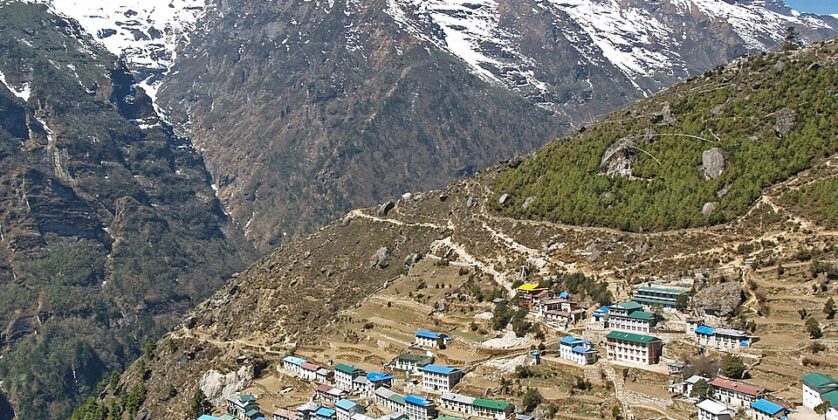 Namche Bazaar village in Nepal with colorful rooftops on terraced hillsides and snow-capped Himalayan mountains in the background.