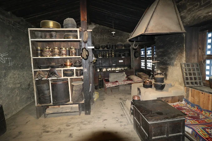 Traditional Sherpa kitchen exhibit in a museum, featuring a stone stove with chimney and wooden racks holding utensils for storing yak milk products, alcohol, and grains.