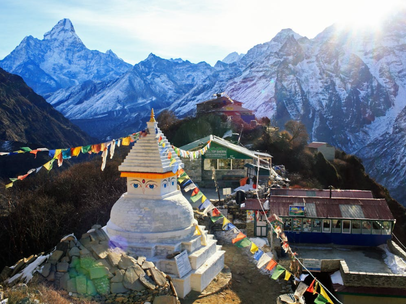 A white Buddhist stupa with painted eyes and colorful prayer flags stands along the Khumbu trekking route, with snow-covered Himalayan peaks rising in the background.
