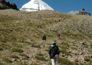 Trekkers hiking up a rocky trail toward the snow-covered Asthapad peak in the Kailash Mansarovar region under a clear blue sky.