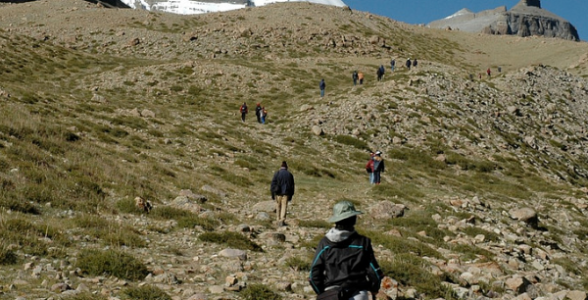 Trekkers hiking up a rocky trail toward the snow-covered Asthapad peak in the Kailash Mansarovar region under a clear blue sky.