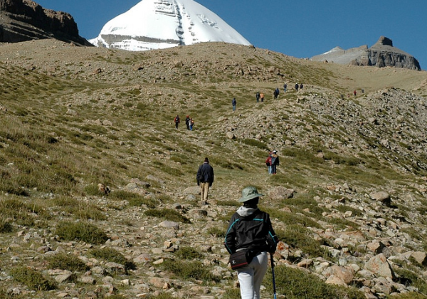 Trekkers hiking up a rocky trail toward the snow-covered Asthapad peak in the Kailash Mansarovar region under a clear blue sky.