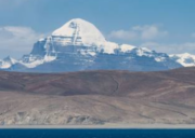 Snow-covered Mount Kailash rising above barren hills with Lake Mansarovar in the foreground under a clear blue sky