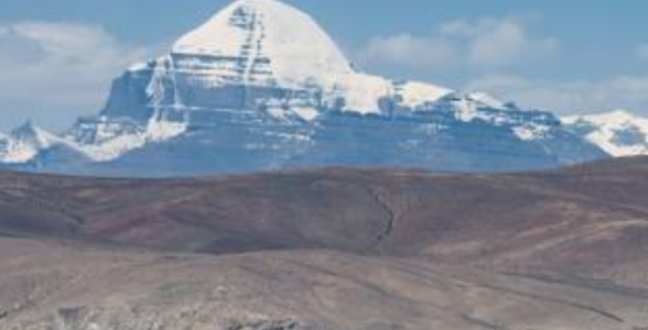 Snow-covered Mount Kailash rising above barren hills with Lake Mansarovar in the foreground under a clear blue sky