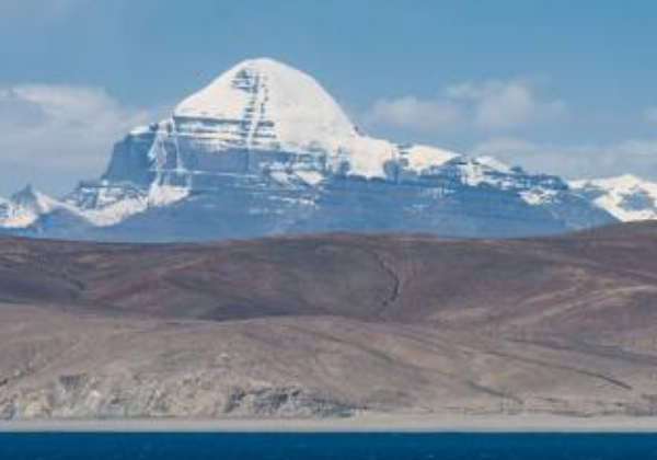Snow-covered Mount Kailash rising above barren hills with Lake Mansarovar in the foreground under a clear blue sky