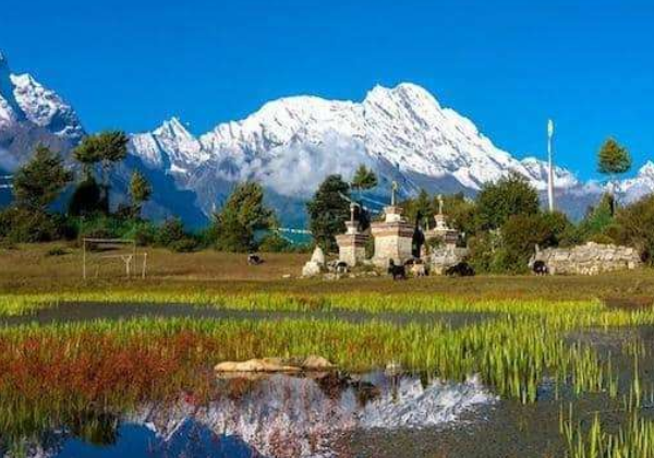 Lush green fields and small stone shrines in Kerung (Kyirong) with snow-capped Himalayan peaks reflected in a calm water pool.