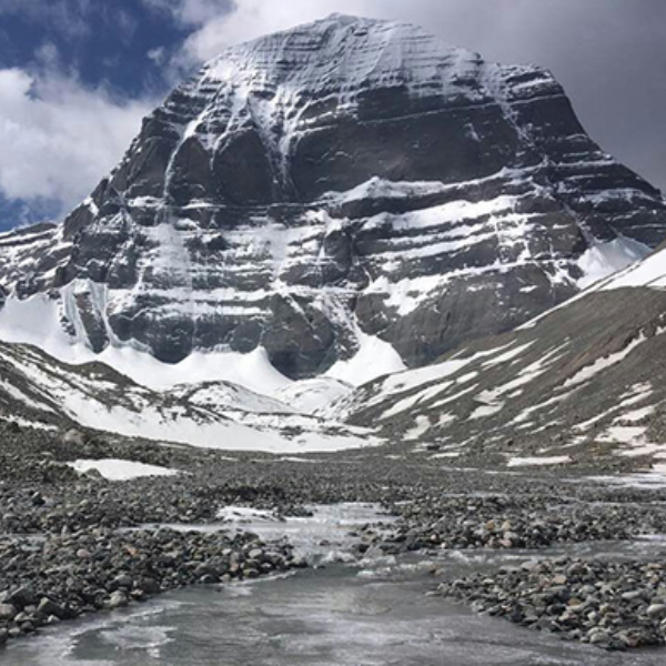 Stream flowing from the base of Mount Kailash through a rocky, snow-dusted valley under a partly cloudy sky