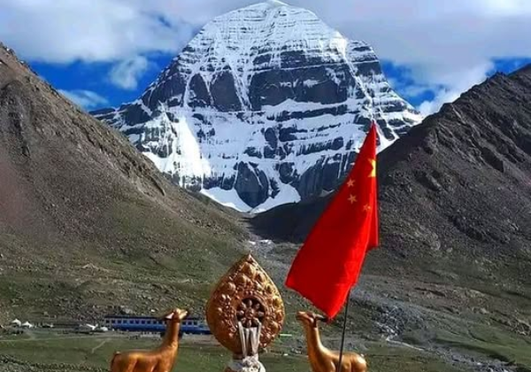 Snow-covered south face of Mount Kailash viewed from DiraPuk Monastery, with a prayer shrine and flags in the foreground against a clear Himalayan sky.