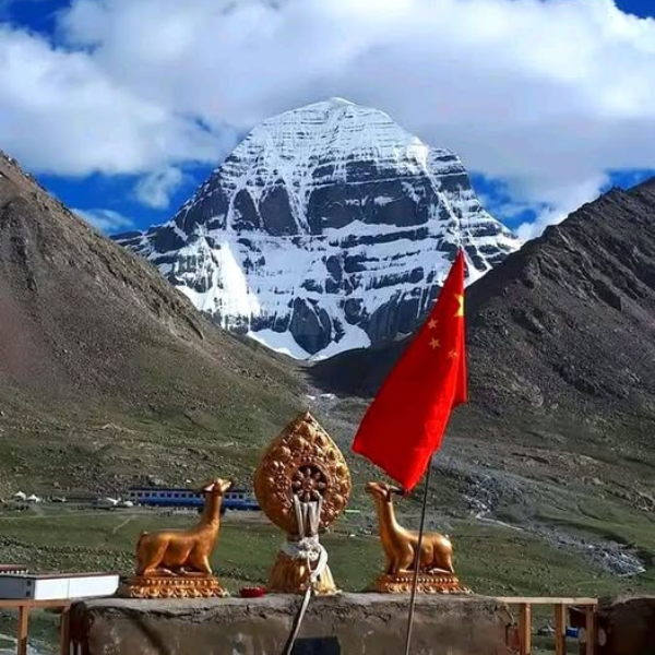 Snow-covered south face of Mount Kailash viewed from DiraPuk Monastery, with a prayer shrine and flags in the foreground against a clear Himalayan sky.
