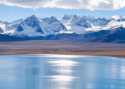 Crystal-clear blue waters of Lake Mansarovar reflecting sunlight, with snow-capped Himalayan mountains and a partly cloudy sky in the background.