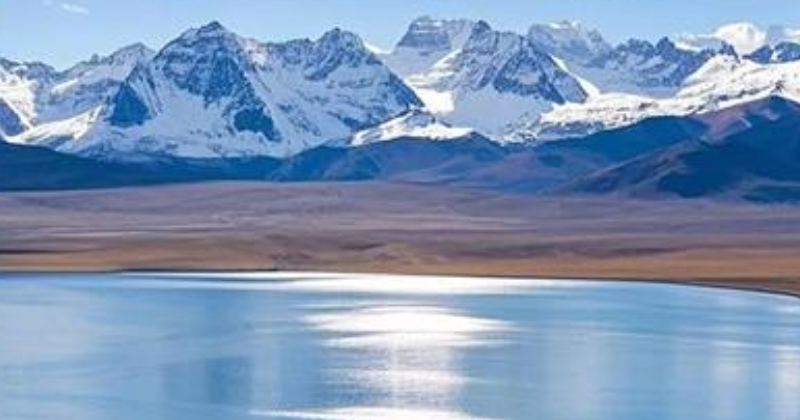Crystal-clear blue waters of Lake Mansarovar reflecting sunlight, with snow-capped Himalayan mountains and a partly cloudy sky in the background.
