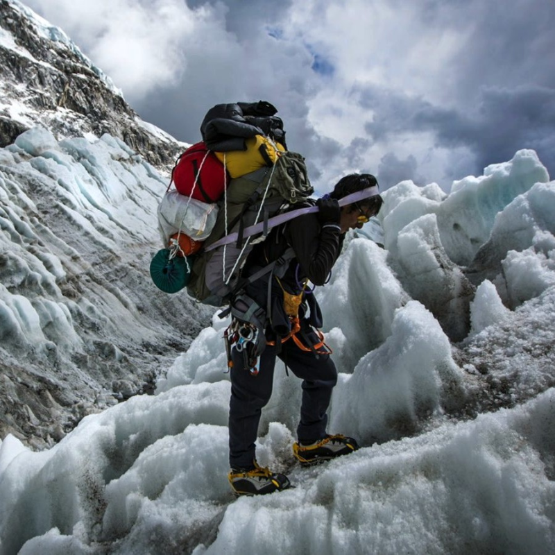 Sherpa mountaineer carrying heavy climbing gear across an icy glacier