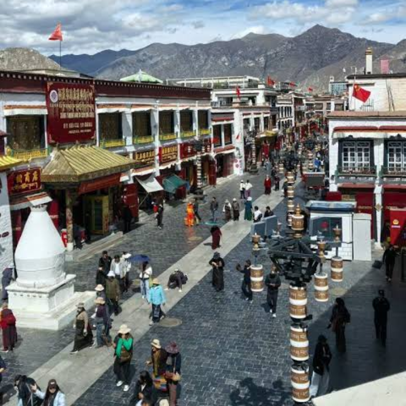 Barkhor Street in Lhasa, Tibet, bustling with pilgrims, shoppers, and visitors walking along the historic market street with traditional Tibetan architecture and mountain views.