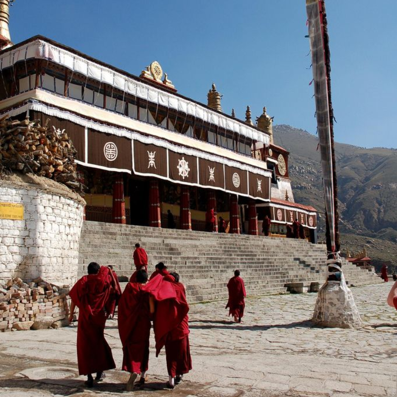 Tibetan Buddhist monks in red robes walking toward the main assembly hall of Drepung Monastery in Lhasa, Tibet, with mountains in the background.