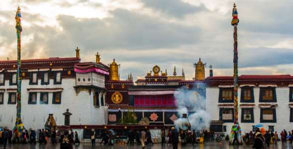 Jokhang Temple in Lhasa, Tibet, with pilgrims and visitors gathered in the courtyard on a rainy day beneath dramatic clouds and rising incense smoke.