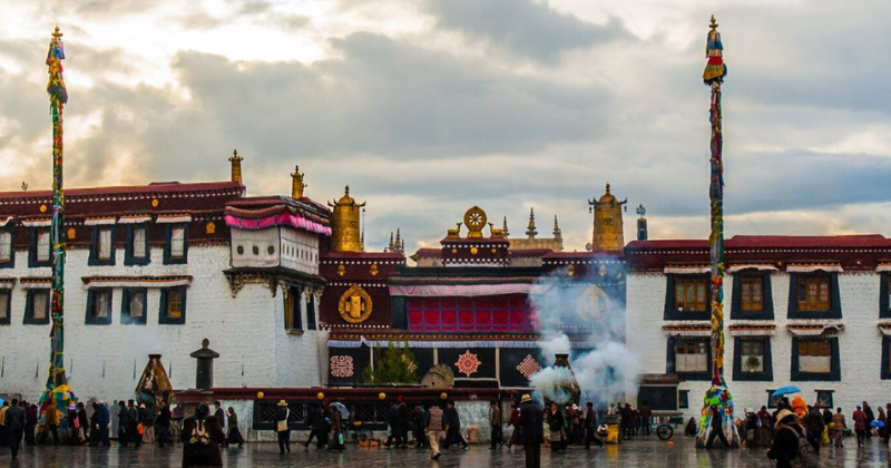 Jokhang Temple in Lhasa, Tibet, with pilgrims and visitors gathered in the courtyard on a rainy day beneath dramatic clouds and rising incense smoke.