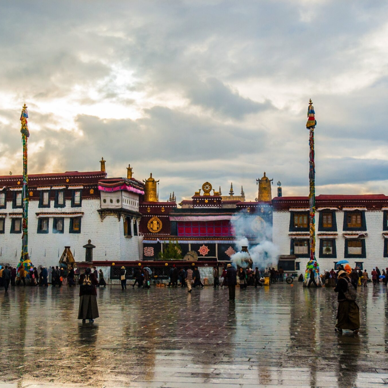 Jokhang Temple in Lhasa, Tibet, with pilgrims and visitors gathered in the courtyard on a rainy day beneath dramatic clouds and rising incense smoke.