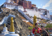 Potala Palace towering above Lhasa, Tibet, with visitors in traditional Tibetan clothing standing in the foreground against the historic white and red palace walls.