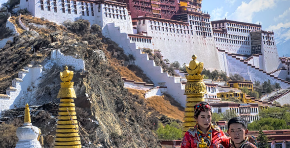 Potala Palace towering above Lhasa, Tibet, with visitors in traditional Tibetan clothing standing in the foreground against the historic white and red palace walls.