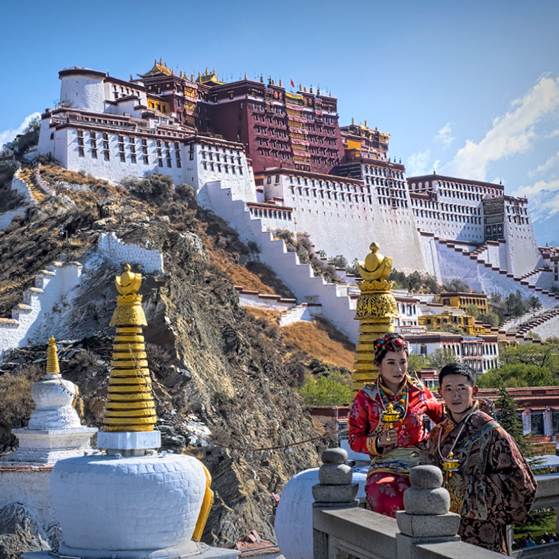 Potala Palace towering above Lhasa, Tibet, with visitors in traditional Tibetan clothing standing in the foreground against the historic white and red palace walls.